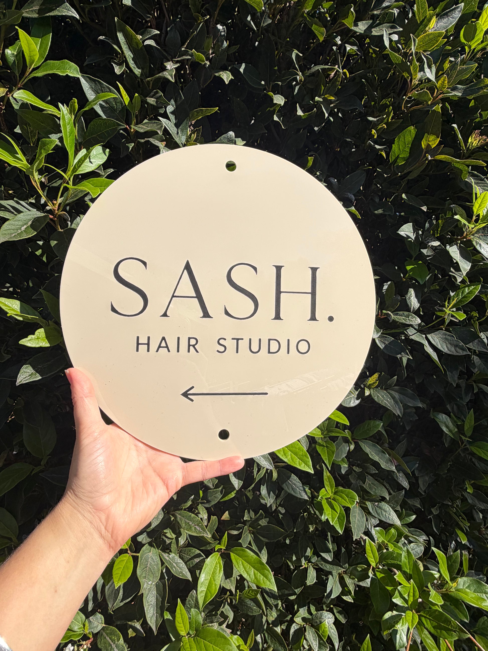 Hand holding a sign for SASH. Hair Studio against a green leafy background