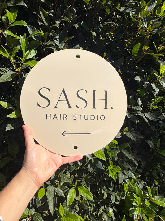 Hand holding a sign for SASH. Hair Studio against a green leafy background
